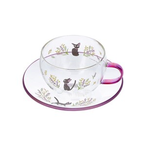 Op de afbeelding: Een theekop en schotel van helder glas met een delicaat bloemmotief van gele en paarse takjes, afgewisseld met kleine illustraties van zwarte katten. De kop heeft een handvat met een roze en paars kleurverloop en een paarse rand.