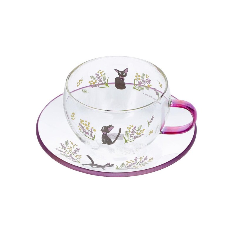 Op de afbeelding: Een theekop en schotel van helder glas met een delicaat bloemmotief van gele en paarse takjes, afgewisseld met kleine illustraties van zwarte katten. De kop heeft een handvat met een roze en paars kleurverloop en een paarse rand.