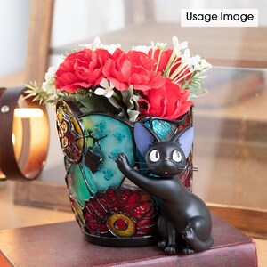 K&ouml;nnte beinhalten: Eine farbige Glasvase mit einer schwarzen Katzenfigur. Die Vase hat ein buntes Design mit roten, gelben, blauen und gr&uuml;nen Glasst&uuml;cken. Die Katze sitzt auf der Vase und hat einen schwarzen K&ouml;rper mit blauen Ohren. Die Vase ist mit roten und wei&szlig;en Blumen gef&uuml;llt.
