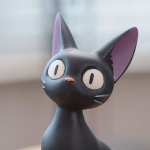 May include: A black cat figurine with large, oval white eyes and a small pink nose. The cat has large, pointed ears with purple insides and white whiskers. The figurine is a stylized, cartoon-like representation of a cat.