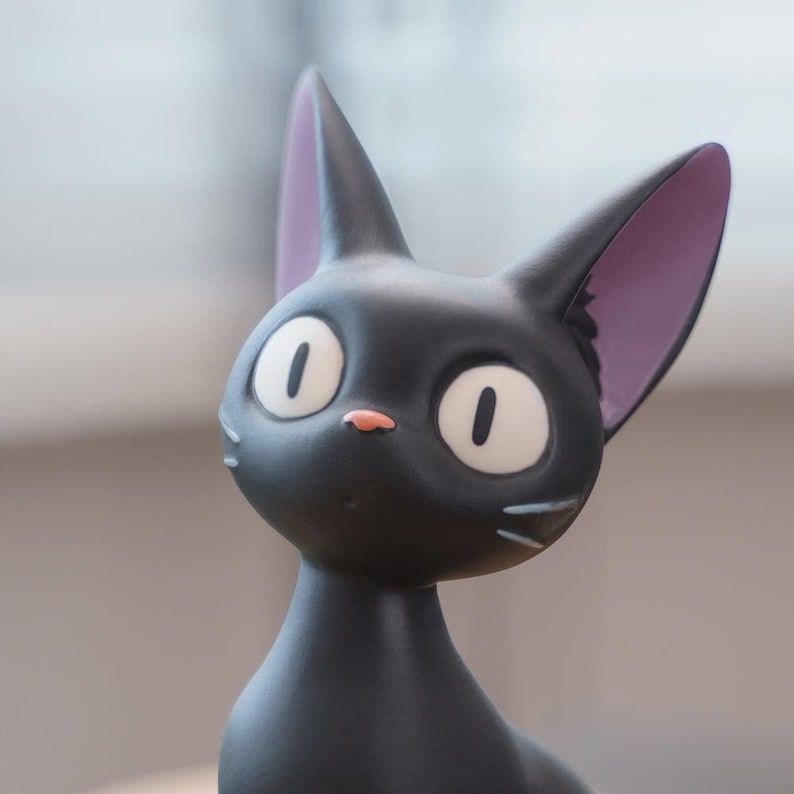May include: A black cat figurine with large, oval white eyes and a small pink nose. The cat has large, pointed ears with purple insides and white whiskers. The figurine is a stylized, cartoon-like representation of a cat.