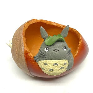 K&ouml;nnte beinhalten: Eine Figur von Totoro, einem grauen Waldgeist, blickt aus einer gro&szlig;en, ausgeh&ouml;hlten Eichel hervor. Die Eichel ist braun und rot, mit glatter Oberfl&auml;che. Totoro tr&auml;gt ein gr&uuml;nes Blatt auf dem Kopf und hat einen cremefarbenen Bauch.