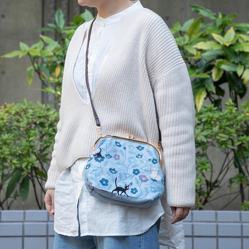 May include: A light blue crossbody bag with a floral and cat pattern. The bag has a gold-colored clasp and a brown strap. The design includes black cats, blue flowers, and purple accents. The bag is worn over the shoulder.
