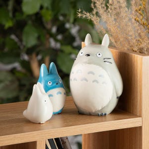 May include: Three ceramic Totoro figurines in varying sizes. The largest is gray with a white belly and gray markings. The medium-sized figurine is blue and white, and the smallest is white. They are displayed on a wooden shelf.