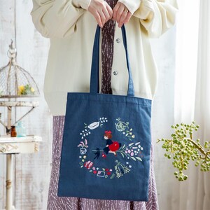 K&ouml;nnte beinhalten: Eine blaue Stofftasche mit einem bunten Stickmuster, das ein M&auml;dchen auf einem Besen mit einer schwarzen Katze zeigt. Das Design enth&auml;lt Blumen, Bl&auml;tter und andere fantasievolle Details.
