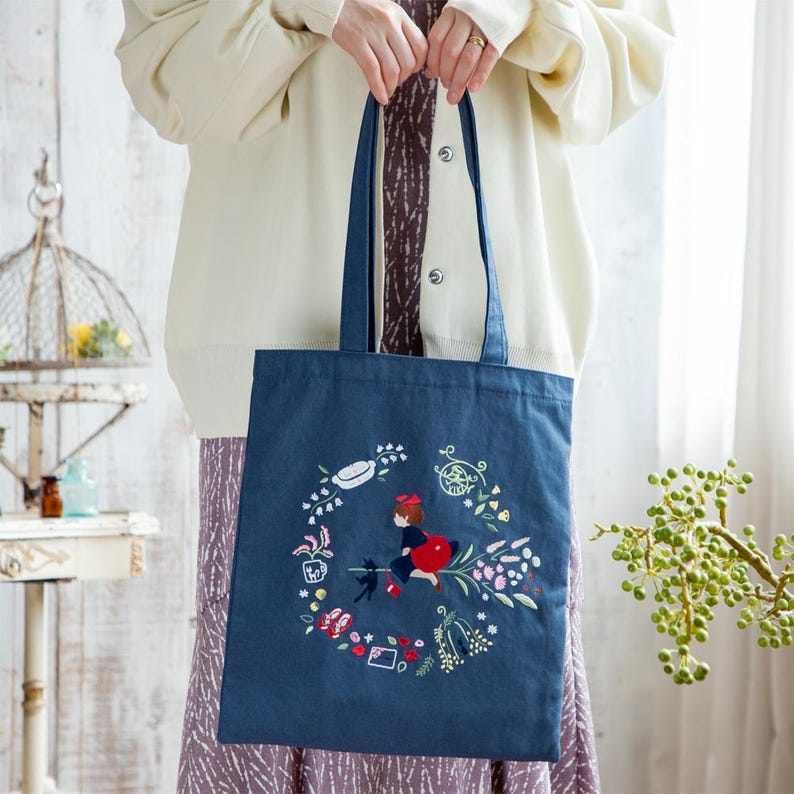 K&ouml;nnte beinhalten: Eine blaue Stofftasche mit einem bunten Stickmuster, das ein M&auml;dchen auf einem Besen mit einer schwarzen Katze zeigt. Das Design enth&auml;lt Blumen, Bl&auml;tter und andere fantasievolle Details.
