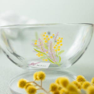May include: A clear glass bowl featuring a printed design of yellow mimosa flowers and light purple lavender sprigs. A small white rectangular label with black text is attached to the side of the bowl, which sits above yellow flowers.