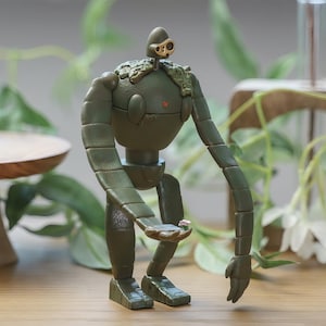 Peut inclure: Figurine d&eacute;taill&eacute;e d'un robot vert, tenant une petite fleur rose. Le robot a des membres segment&eacute;s et une t&ecirc;te avec un d&eacute;tail en forme d'&oelig;il. L'ensemble sugg&egrave;re un th&egrave;me fantastique ou anim&eacute;. La figurine est tr&egrave;s d&eacute;taill&eacute;e.