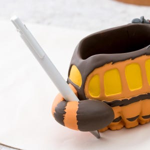 May include: A pen holder shaped like a cartoon bus, featuring a brown and orange color scheme with yellow window details. A white pen is inserted into the holder, showcasing its functionality. The design is whimsical and suitable for desk organization.