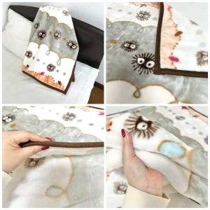 Peut inclure: Une couverture en peluche douce avec un motif r&eacute;p&eacute;t&eacute; de sprites de suie de dessin anim&eacute; sur un motif de nuages gris et blancs. La couverture a une bordure marron et est pli&eacute;e sur un lit.