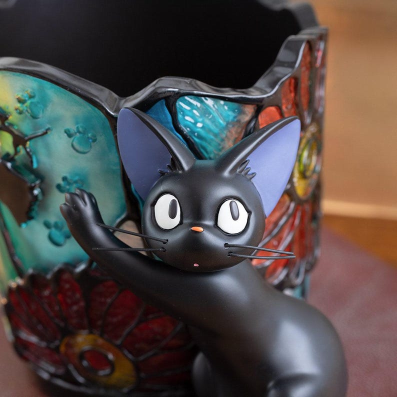 K&ouml;nnte beinhalten: Eine schwarze Katzenfigur mit gro&szlig;en, runden Augen und einem &uuml;berraschten Ausdruck. Die Katze sitzt auf einem bunten, mosaikartigen Sockel mit floralen Designs.