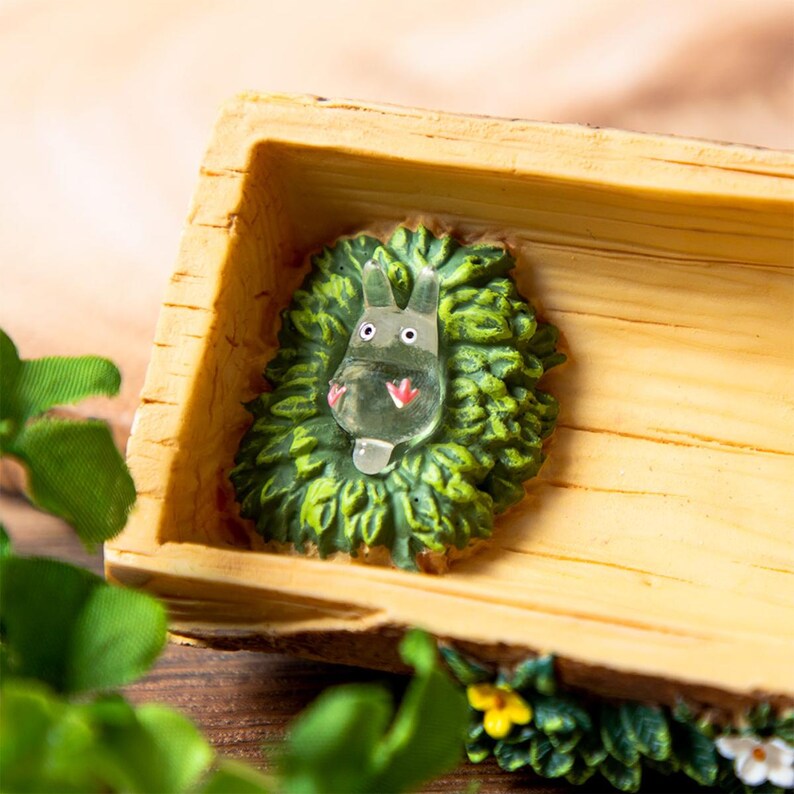 May include: A decorative wooden box with a carved, light-colored exterior. Inside, a green, leafy base holds a clear, cartoon-like figure with a red nose and white eyes. The box is decorated with small flowers and greenery.