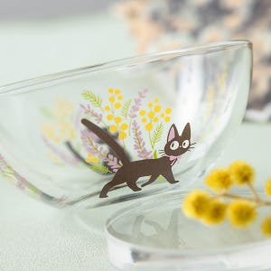 May include: A clear glass bowl featuring a printed graphic of a black cat with a pink collar, surrounded by yellow mimosa flowers and purple lavender sprigs. The bowl has a smooth, curved rim and a transparent body that displays the floral design.