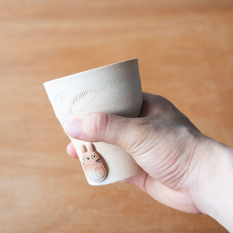 May include: A handmade, off-white ceramic cup with a textured surface. The cup features a carved leaf design and a small, brown Totoro figure attached to the side. The cup is held in a hand, showcasing its size and shape.