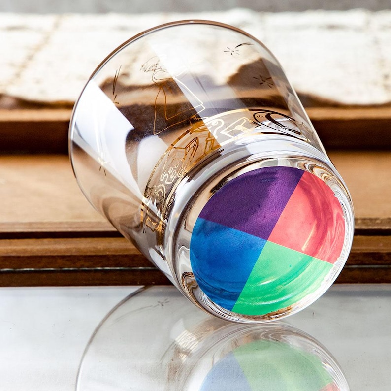 May include: A clear glass tumbler with a colorful, segmented design on the bottom. The glass has gold-colored decorative accents around the rim. The bottom of the glass is divided into four sections of blue, green, red, and purple.