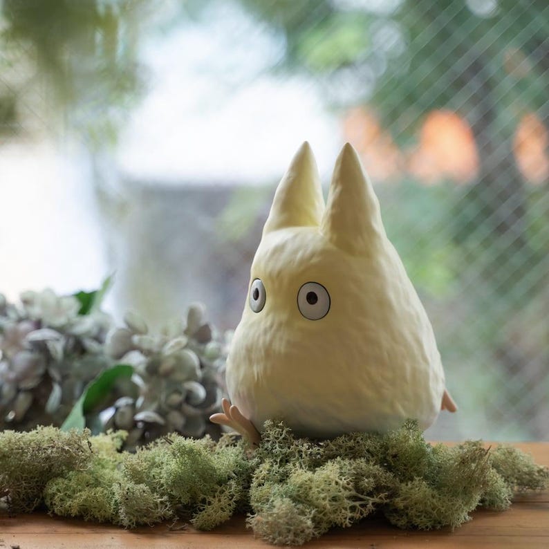 May include: A plush, light yellow Totoro character with large, round eyes and pointed ears. The stuffed animal is sitting on a bed of green moss, with a blurred background of greenery and a window.