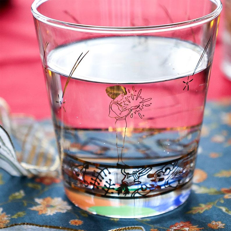 May include: A clear glass tumbler filled with pink liquid. The glass features a decorative illustration of a person holding a glowing object. The base of the glass has a colorful, ornate design. The glass is on a floral patterned surface.