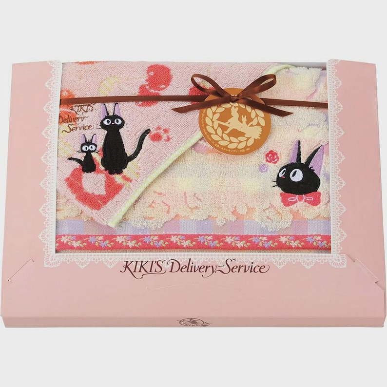 May include: A pink gift box featuring a Kiki's Delivery Service themed towel set. The set includes a towel with embroidered black cat characters and a decorative ribbon. The box has the text "KIKIS Delivery Service".