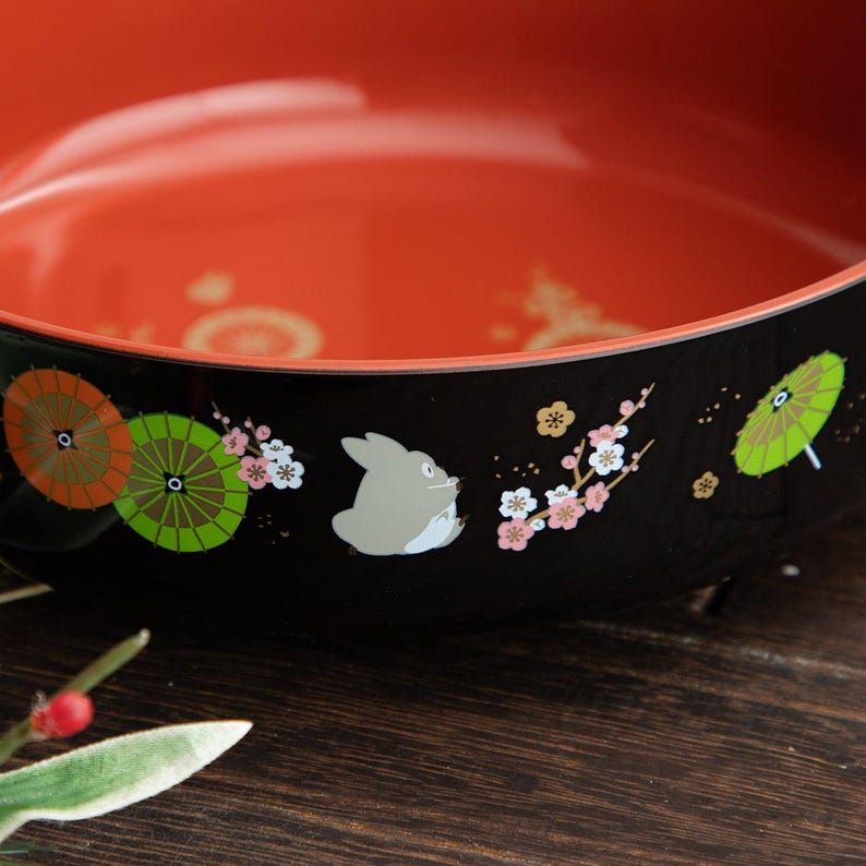 May include: A black and red decorative bowl with a cartoon illustration of a gray rabbit-like creature, umbrellas, and cherry blossoms. The bowl has a glossy finish and is likely used for serving food or as a decorative piece.