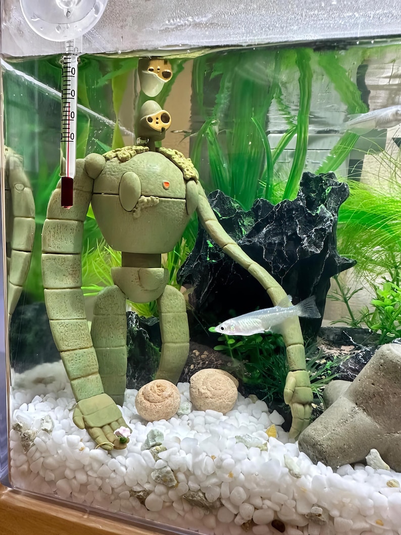 Peut inclure: Un aquarium avec une figurine de robot vert clair, un thermom&egrave;tre et un petit poisson. Le r&eacute;servoir contient du gravier blanc, des plantes vertes et une roche noire. Le robot a un design d&eacute;taill&eacute; avec des membres segment&eacute;s et une t&ecirc;te avec plusieurs yeux.