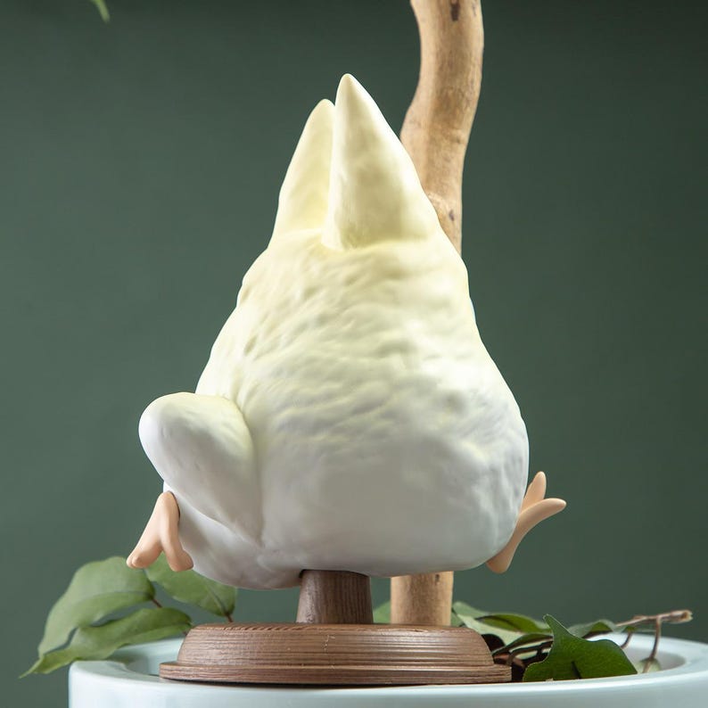 May include: A ceramic figurine of a bird, primarily white with yellow highlights, perched upside down on a wooden stand. The bird's tail feathers and feet are visible. The background is a muted green.