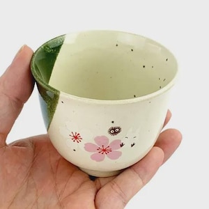 May include: A ceramic teacup with a cream-colored exterior, adorned with pink cherry blossom designs and small black speckles. The rim is green. The teacup is held in a hand.