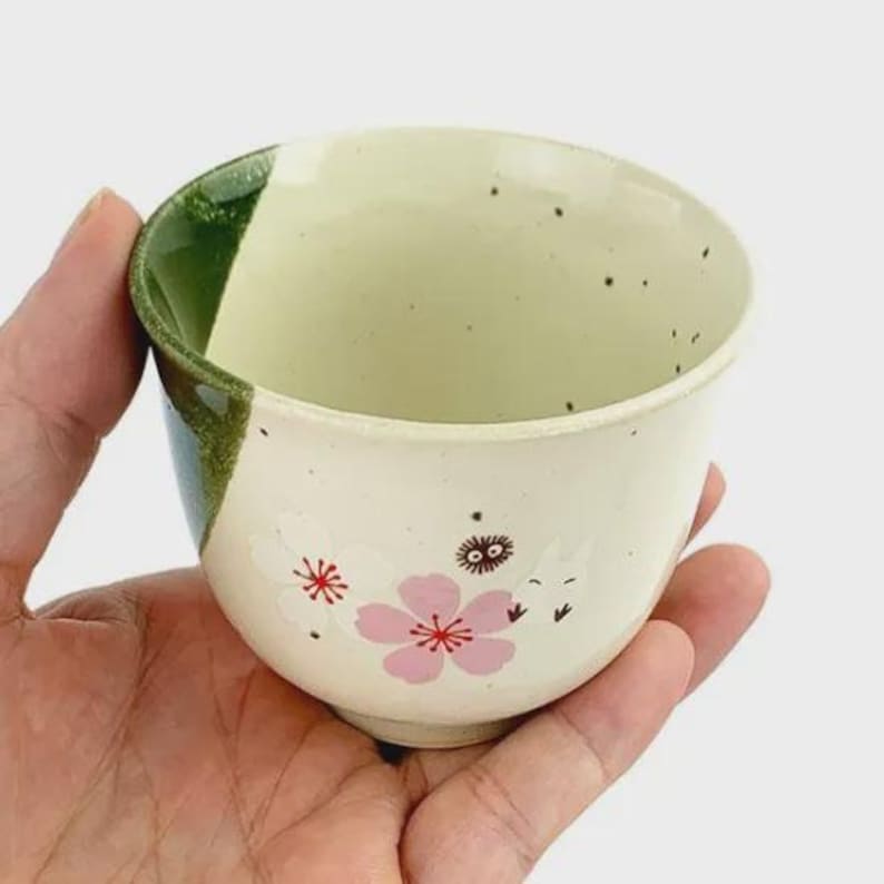May include: A ceramic teacup with a cream-colored exterior, adorned with pink cherry blossom designs and small black speckles. The rim is green. The teacup is held in a hand.