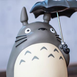 Puede incluir: Una figura gris de Totoro con orejas puntiagudas y bigotes negros, que presenta un vientre de color crema con marcas de cheur&oacute;n gris oscuro. El personaje sostiene un paraguas negro con mango plateado sobre su cabeza, mostrando grandes ojos blancos.