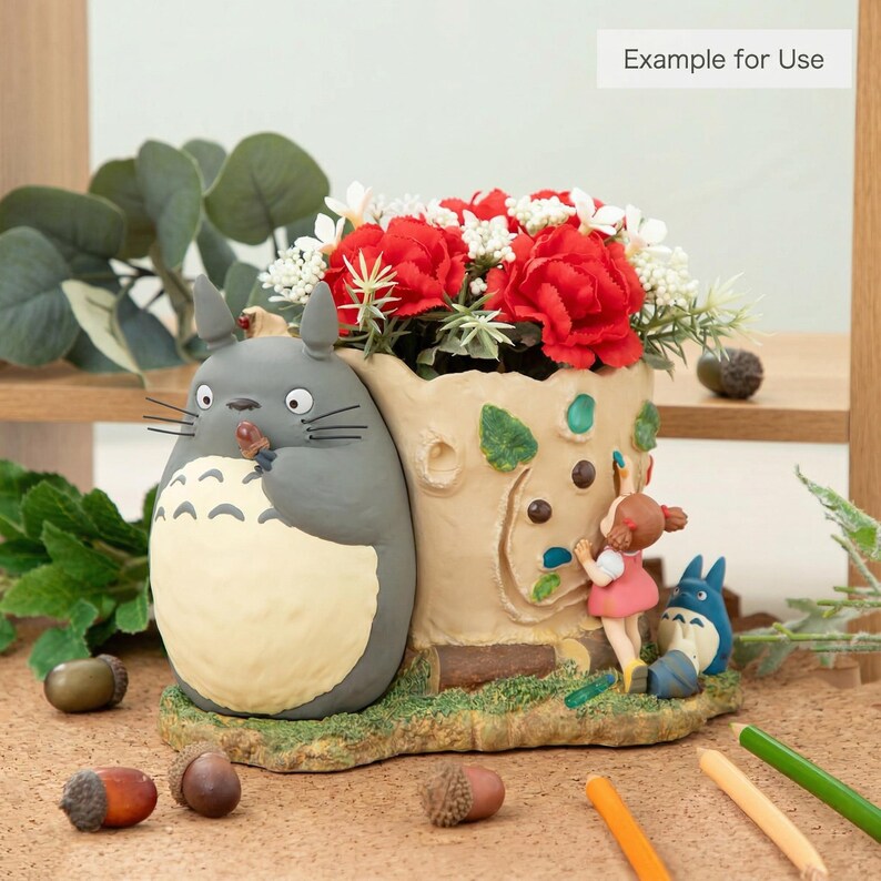 Puede incluir: Una maceta decorativa con una figura de Totoro gris sosteniendo una nuez, con una maceta en forma de tronco de &aacute;rbol llena de flores rojas. Tambi&eacute;n hay una ni&ntilde;a y una figura de Totoro azul. La maceta est&aacute; sobre una base marr&oacute;n con bellotas.