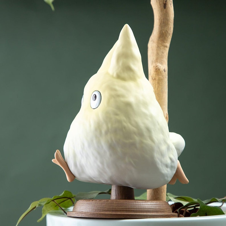 May include: A whimsical, ceramic bird figurine in soft yellow, perched on a wooden stand with a branch. The bird has a rounded body, a pointed head, and a single visible eye. The figurine is set against a green background with foliage.