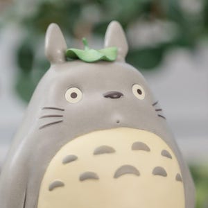 May include: A gray figurine of a cartoon character with large eyes, a small nose, and whiskers. The character has a light yellow belly with dark gray spots and a green leaf on its head. The figurine is a collectible item.