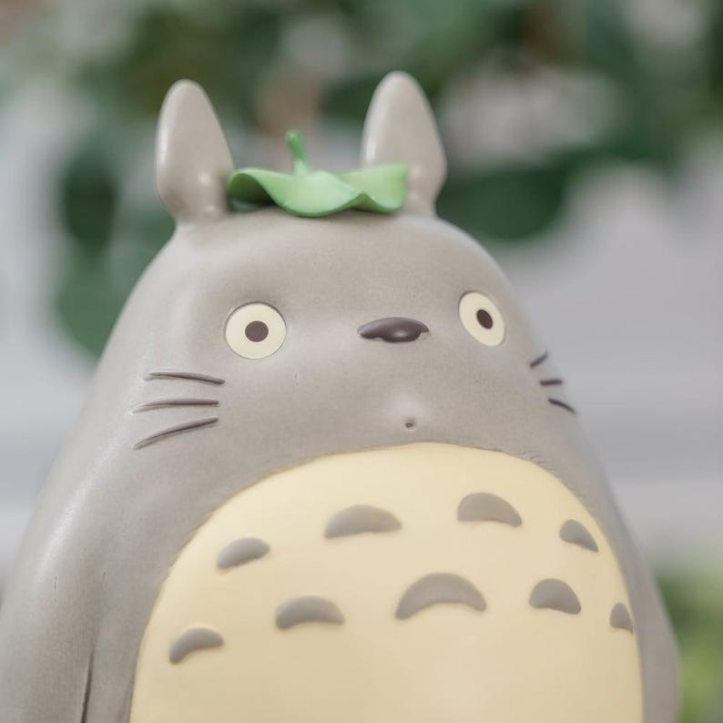 May include: A gray figurine of a cartoon character with large eyes, a small nose, and whiskers. The character has a light yellow belly with dark gray spots and a green leaf on its head. The figurine is a collectible item.