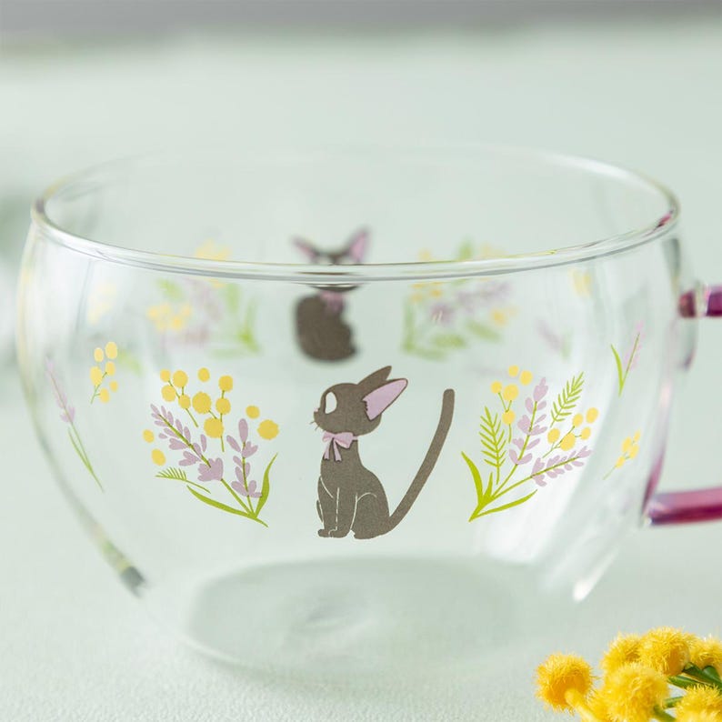 Op de afbeelding: Een doorzichtige glazen theekop met een herhalend patroon van een kleine zwarte kat met een lichtroze strik om de nek, afgewisseld met illustraties van gele mimosa bloemen en takjes van lichtpaarse lavendel, met een paars handvat.