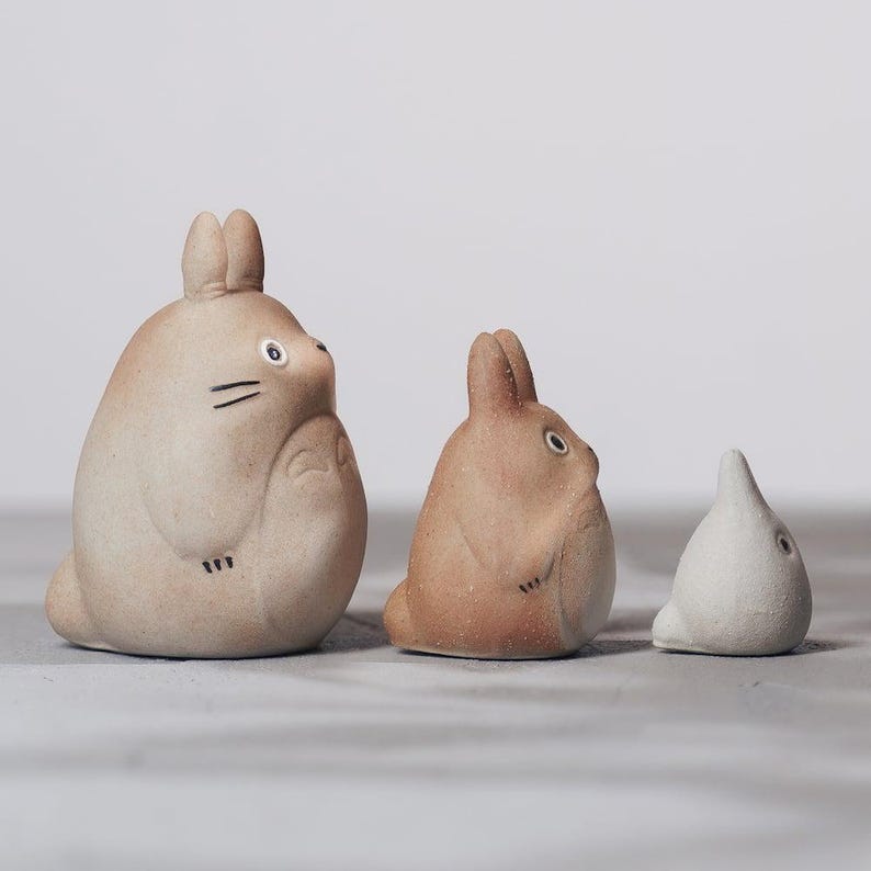 May include: Three ceramic figurines in varying sizes and colors. The largest is tan with black details, the middle is brown, and the smallest is white. All have a rounded shape with bunny-like ears and simple facial features.