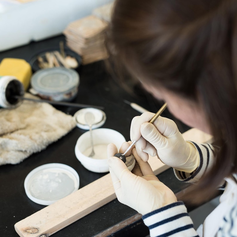 Puede incluir: Una persona con guantes de l&aacute;tex blancos utiliza un pincel fino y puntiagudo para aplicar pintura sobre un peque&ntilde;o objeto esf&eacute;rico oscuro. El espacio de trabajo incluye varios cuencos de cer&aacute;mica blanca con l&iacute;quidos, una tabla de madera y utensilios.