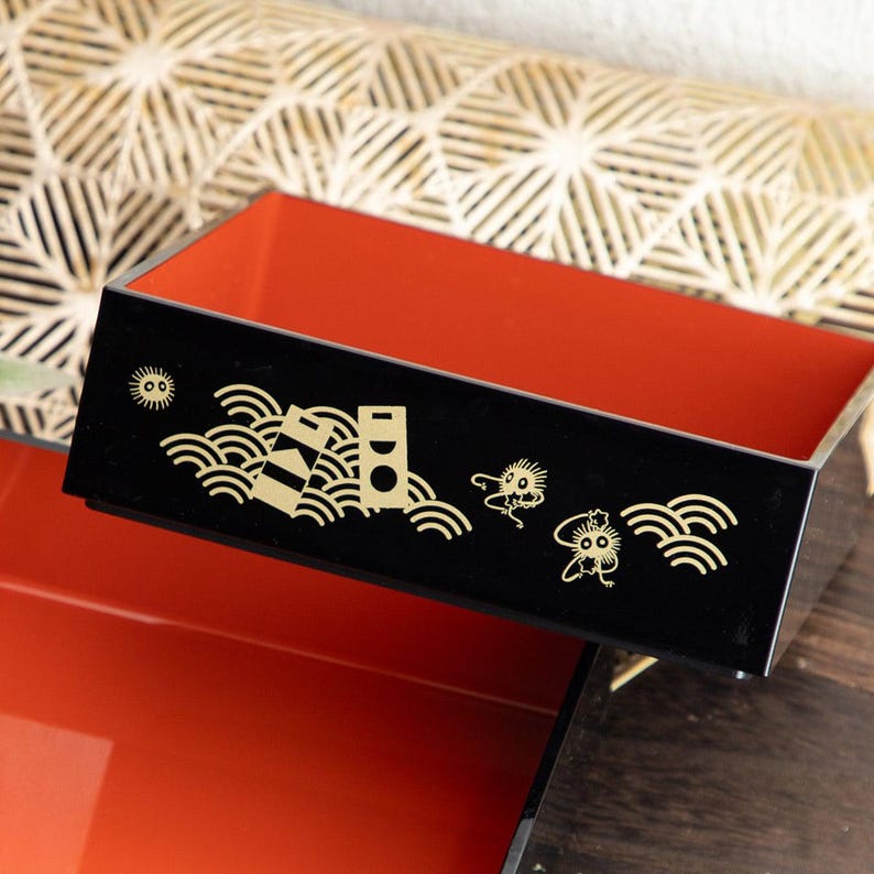 May include: A black lacquered bento box with a red interior. The box is decorated with gold illustrations of waves, suns, and whimsical creatures. The box is rectangular and has a lid.