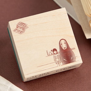 May include: A square wooden rubber stamp featuring a brown ink illustration of two characters, one tall and hooded and one smaller, with a small stamp-like graphic in the top left corner and Japanese text printed along the bottom edge.