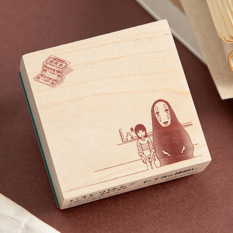May include: A square wooden rubber stamp featuring a brown ink illustration of two characters, one tall and hooded and one smaller, with a small stamp-like graphic in the top left corner and Japanese text printed along the bottom edge.