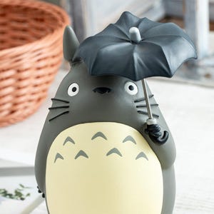 Puede incluir: Una figura de criatura gris y redondeada con orejas puntiagudas, bigotes negros y una barriga color crema con marcas de chevr&oacute;n gris oscuro. La figura sostiene un paraguas gris oscuro con un mango plateado y una peque&ntilde;a punta blanca.