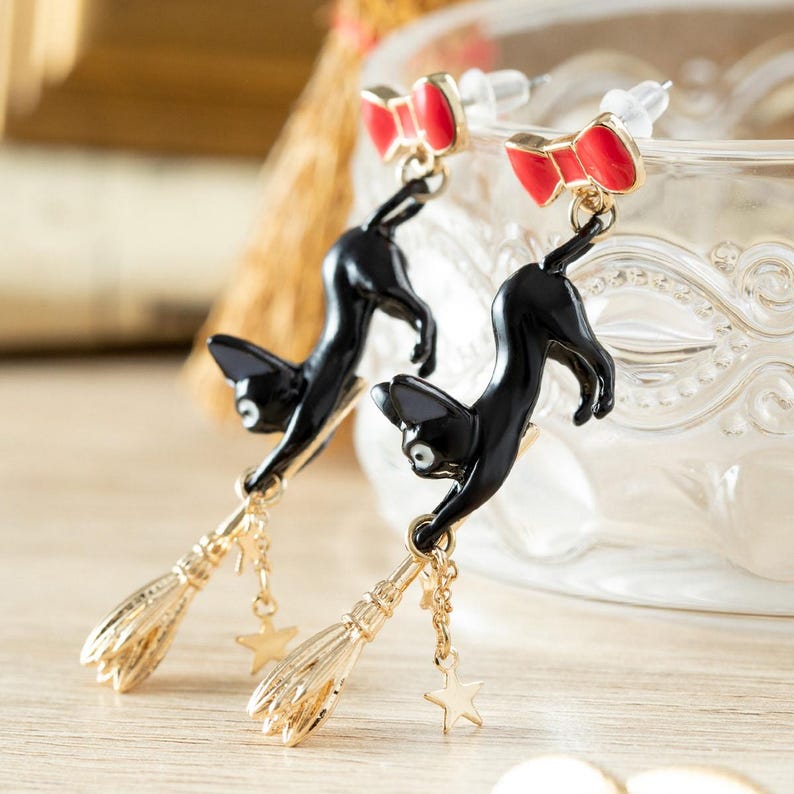 May include: A pair of black cat earrings with red bow accents. The earrings feature a gold-colored broom and star charm. The cat is in a playful pose, hanging from the broom. The earrings are set against a blurred background.
