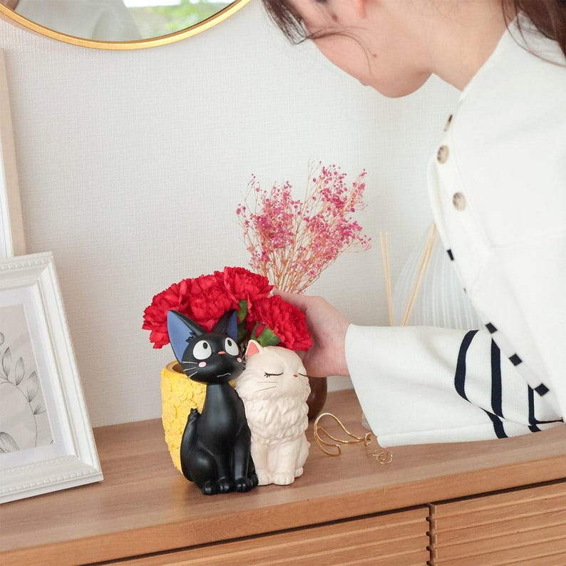 Puede incluir: Una maceta decorativa con dos gatos esculpidos, uno negro de ojos grandes y otro blanco de ojos cerrados, sentados uno al lado del otro. El gato negro est&aacute; frente a un recipiente amarillo texturizado que contiene claveles rojos.