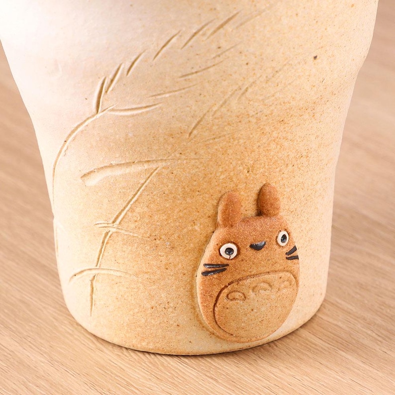May include: A beige ceramic mug with a textured surface and a raised leaf design. A small, brown, cartoon-like character is affixed to the side of the mug. The character has large eyes, a round body, and small ears.