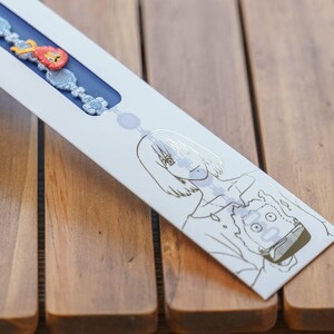 Op de afbeelding: Een witte productverpakking met een blauw interieur, met een armband met rode, oranje en blauwe decoratieve elementen. De verpakking heeft gouden lijntekeningen van twee anime-achtige personages. De achtergrond is een houten oppervlak.