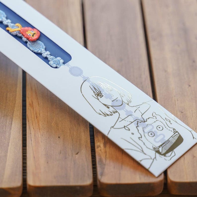 Op de afbeelding: Een witte productverpakking met een blauw interieur, met een armband met rode, oranje en blauwe decoratieve elementen. De verpakking heeft gouden lijntekeningen van twee anime-achtige personages. De achtergrond is een houten oppervlak.