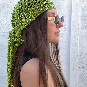 Puede incluir: Un gorro verde tejido a ganchillo con lentejuelas doradas y una larga cola fluida. El gorro se lleva con gafas de sol con montura dorada. El fondo es una pared blanca y una valla de madera.