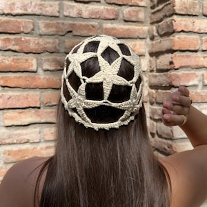 Puede incluir: Un sombrero de ganchillo color crema con un patrón de estrellas. El sombrero se lleva en la cabeza de una persona, con pelo largo castaño visible. El fondo es una pared de ladrillo.