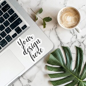 Könnte beinhalten: Ein silberner Laptop mit einem weißen Aufkleber, auf dem "Your design here" steht, auf einer weißen Marmorfläche mit einem grünen Blatt und einer weißen Tasse Kaffee mit Latte Art.