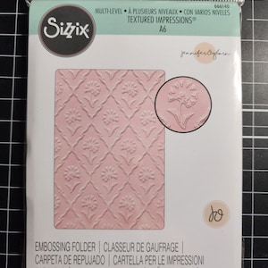Puede incluir: Una carpeta de repujado Sizzix A6 Multi-Level Textured Impressions en un empaque de plástico transparente. El producto presenta un patrón floral rosa claro con un diseño de celosía repetitivo e incluye un inserto circular con el detalle.