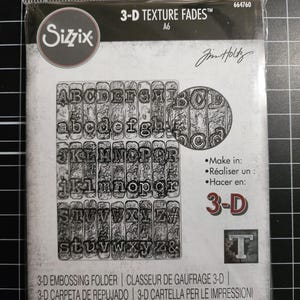 Op de afbeelding: Een Sizzix 3-D Texture Fades A6 embossing folder in een doorzichtige plastic verpakking. Het product bevat een alfabetontwerp met hoofdletters en kleine letters op een getextureerde achtergrond. De verpakking bevat de tekst 3-D Texture Fades, Sizzix, Tim Holtz en 664760.