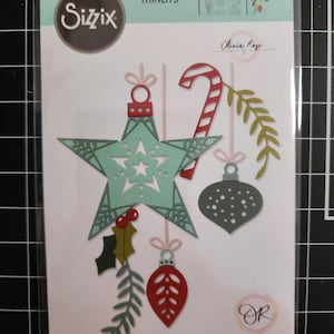 May include: Sizzix Thinlits die set 666685 by Olivia Rose featuring 15 metal cutting dies. The packaging displays die-cut shapes including a light green star ornament, a red and white candy cane, a grey bauble, red berries, and green foliage.